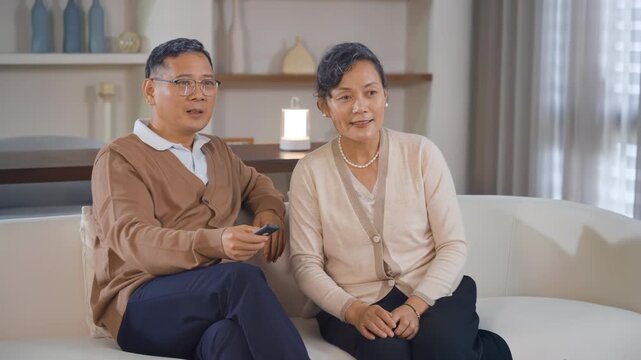 Happy elderly couple wife husband laughing and watching tv together in retirement, talking holding remote control. Television, smile and elderly man and woman family lifestyle leisure concept