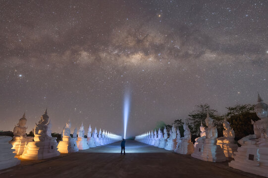 Rear view silhouette of a man holding a torch standing in the middle of a path lined with white Buddha statues at night, Wat Phra That Choeng Chum Worawihan, Sakon Nakhon, Thailand