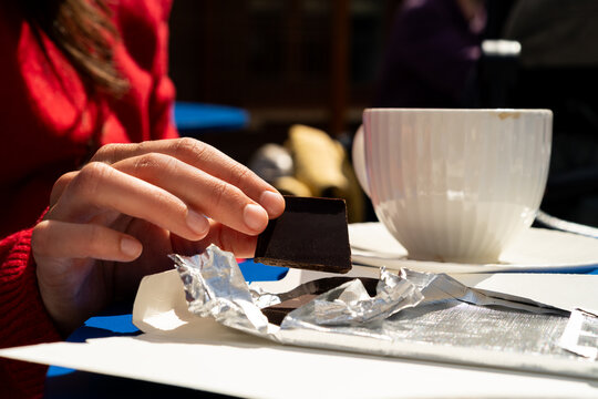Close-up side view of a Woman sitting at an outdoor table enjoying a piece of dark chocolate with a cup of coffee