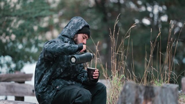 A man in a hooded jacket pours and drinks mate while sitting in a forest clearing. Ideal for outdoor lifestyle and travel content.