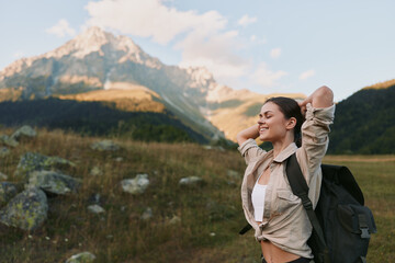 Naklejka premium Cheerful woman outdoors, smiling with sunlit cheeks, hiking with backpack, casual jacket, amidst meadow and mountain scenery, embracing freedom and nature.