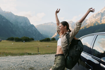 Naklejka premium Smiling woman with a backpack beside a car in a mountain valley, embracing outdoor life, nature escape, and wellness. Bright, airy moment with adventurous spirit. Casual outdoor vibes with travel