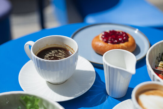 Close-up of a cup of black coffee, cappuccino, cherry brioche bun and assorted plates on a table at an outdoor cafe on a sunny day