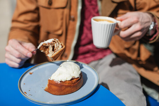 Close-up of a man sitting at an outdoor cafe enjoying an iced cinnamon bun and cappuccino