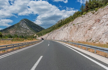 Fototapeta premium An empty asphalt highway curves through a mountainous landscape toward a tunnel under a bright blue sky with white clouds.