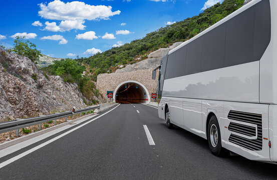 A large white tour bus travels on an asphalt highway toward a stone tunnel entrance under a bright blue sky.