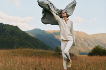 Naklejka premium Woman dances in a field near mountains, wearing white sweater and pants, scarf billowing, carefree lifestyle shot capturing joy, movement and outdoor freedom