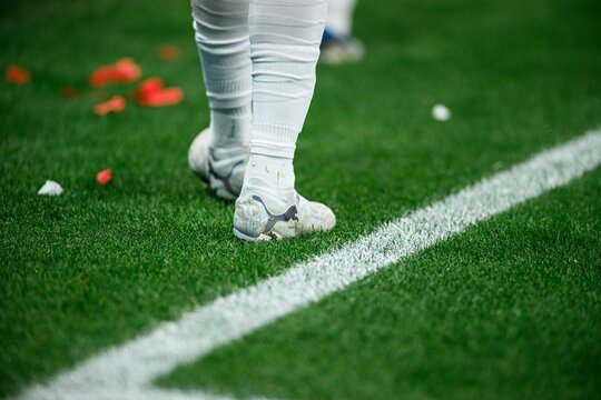 Lower body of a football player walking on the pitch, taped calves and cleats on wet grass during a professional match.