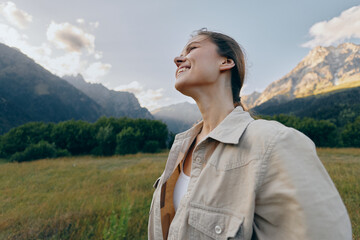 Naklejka premium Cheerful woman outdoors, smiling with eyes closed, sunlit moment in a grassy field, mountains in the background, relaxed posture, natural colors, lifestyle vibe