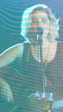 Vertical video: Female musician moving to mic performing song and strumming guitar under teal bands