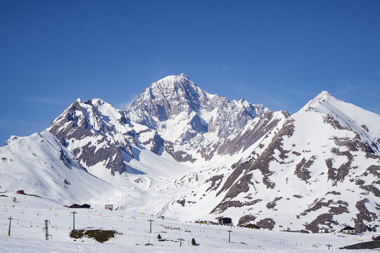 Aerial view of the snow-covered peaks of the Mont Blanc massif and ski slopes with lift infrastructure under a clear blue sky in La Thuile, Valle d'Aosta, Italy.