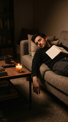 Fototapeta premium Man sleeping on couch next to burning candle. Fire hazard risk due to unattended open flame near flammable objects in cozy room. Negligence of home fire safety and accident prevention.