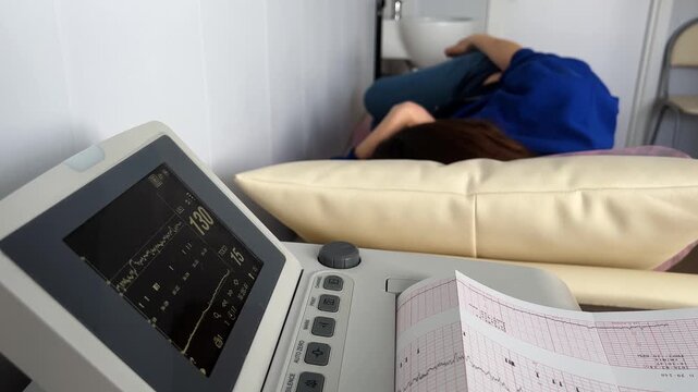 Expectant mother lying on a bed in a medical clinic during a fetal heart rate monitoring examination, with the cardiotocograph machine displaying data and printing a cardiogram