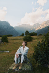 Naklejka premium Woman in a cozy shawl sits on a rock in a mountain meadow, enjoying tranquil nature and a relaxed casual lifestyle portrait amid open sky and distant peaks