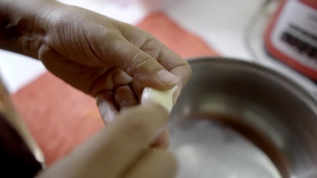 Close view of hand peeling garlic clove over metal bowl, fresh ingredient being prepared for cooking, simple kitchen setting with natural food preparation moment