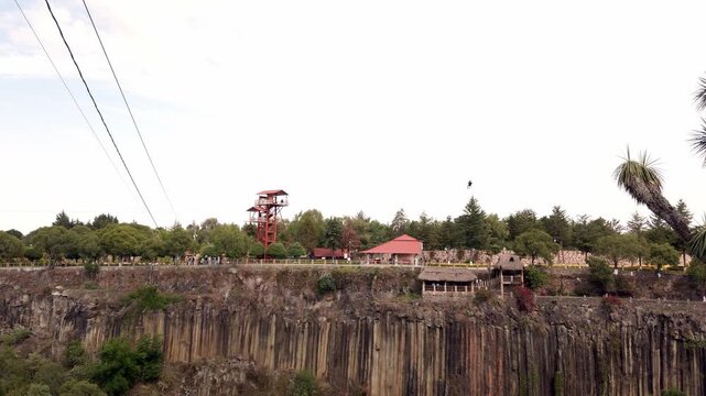 Person ziplining across basalt columns canyon, cables stretching over rock formations with observation tower and trees, outdoor adventure scene in natural landscape