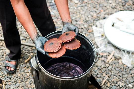 A craftsman with hands stained blue holding molded palm sugar discs over a metal vat containing natural indigo dye mixture.