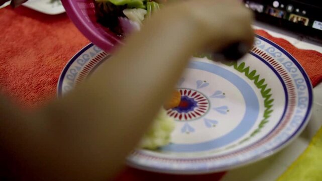 Close view of hand serving broccoli, cauliflower and carrot from plastic strainer onto patterned plate, cooked vegetables placed for meal in simple kitchen setting