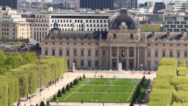The historic &Eacute;cole Militaire building with its distinctive dome, standing behind a lush green lawn and tree-lined pathways of the Champ de Mars, bustling with people enjoying the Parisian landscape