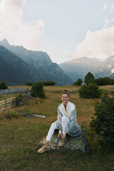 Naklejka premium Casual joyful woman sits on a rock in a scenic mountain meadow, wearing a cardigan and white jeans, smiling, enjoying outdoor life in a peaceful rural landscape under soft daylight.