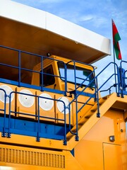 Low angle view of a giant yellow mining dump truck with massive tires, blue metal stairs and safety railings against dramatic cloudy blue sky. Heavy industrial mining vehicle concept. © Алина Неборская