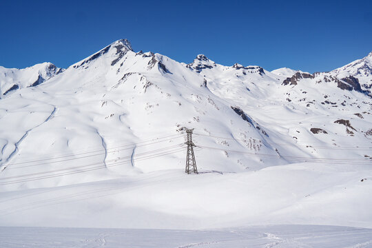 Aerial view of Colle del Piccolo San Bernardo with snow-covered mountain peaks and a high-voltage electricity pylon under a clear blue sky in La Thuile, Valle d'Aosta, Italy.