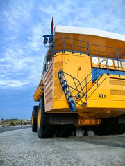 Low angle view of a giant yellow mining dump truck with massive tires, blue metal stairs and safety railings against dramatic cloudy blue sky. Heavy industrial mining vehicle concept. © Алина Неборская