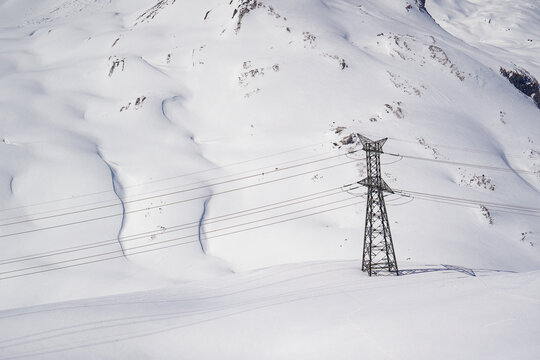 Aerial view of a high-voltage electricity pylon and power lines crossing the snow-covered slopes of Colle del Piccolo San Bernardo in La Thuile, Valle d'Aosta, Italy.