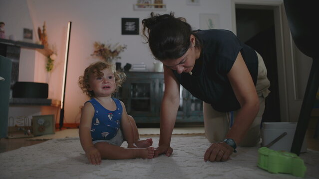 Mother rubbing and cleaning carpet to remove a stain while toddler cries beside her seeking attention, candid indoor family moment showing daily parenting challenges