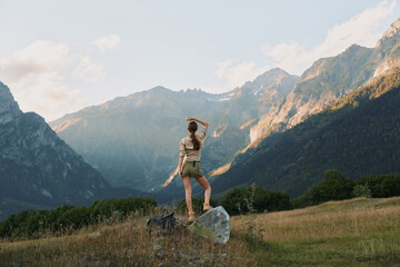 Naklejka premium Woman outdoors in casual attire, enjoying a mountain landscape, daylight sun highlights vibrant nature, wide scenic terrain, relaxed lifestyle moment amid rugged slopes