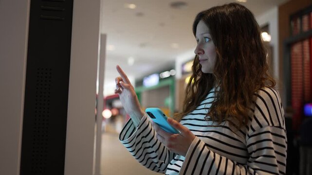 Young expectant mother with curly hair using an interactive touchscreen terminal for ordering at a fast-food restaurant inside a bustling shopping center, holding her smartphone