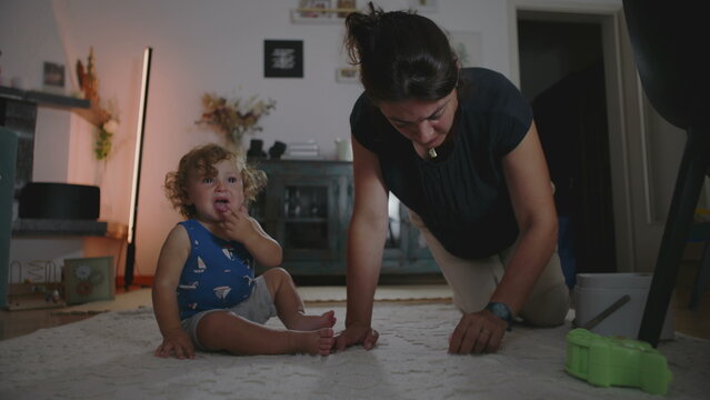 Mother rubbing and cleaning carpet to remove a stain while toddler cries beside her seeking attention, candid indoor family moment showing daily parenting challenges