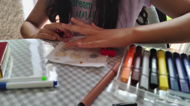 A static close-up high angle video shows a young girl sitting and painting human figures on a sheet of paper. The scene captures her concentration and creativity as she carefully works on her artwork 
