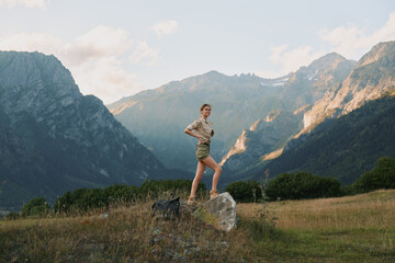 Obraz premium Young woman hiking in mountains stands on rock in open field with tall grass under cloudy sky during daytime, enjoying active lifestyle and nature outdoors.