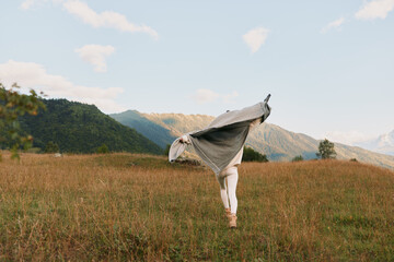 Naklejka premium A woman in a light shawl stretches in a golden meadow with distant mountains, conveying freedom and calm in the open countryside, an airy scene of movement under a bright sky.