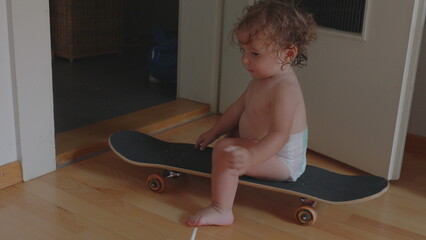Toddler sitting on skateboard wearing diaper indoors, exploring balance and movement, playful childhood moment of curiosity and independence at home © Marco