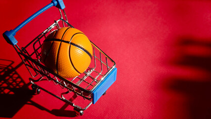 Small shopping cart with an orange basketball inside, placed on a vibrant red background, showcasing a playful and creative concept of sports and shopping