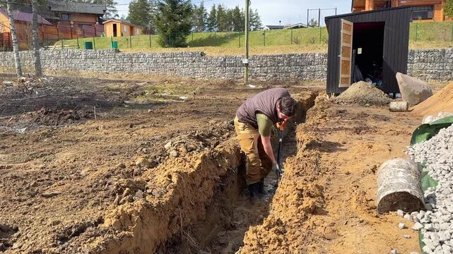 A man digs a drainage trench on his countryside property, working outdoors with a shovel