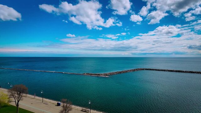 Cinematic aerial drone footage of Marilyn Bell Park along Lake Ontario in Toronto, Canada. The scene captures a vibrant lakeside walking path with pedestrians, light urban traffic, and sweeping views 
