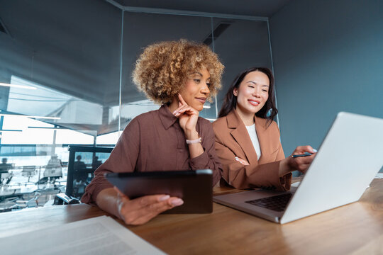 Two professional women collaborating at office desk, reviewing data on laptop and tablet in meeting room, teamwork and business planning. Team of women using technology at work, doing analysing