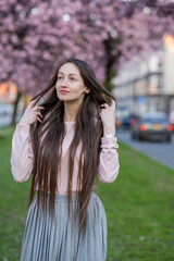 Naklejka premium Woman with long brown hair wearing sunglasses poses among blooming cherry blossom trees in a vibrant spring garden, soft focus background enhancing floral colors