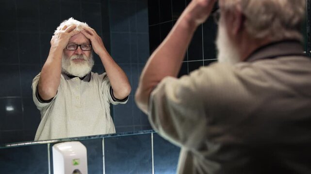 Elderly man in glasses stands in bathroom. He ruffles his hair with his hands
