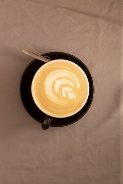 Moody latte with leaf foam on black saucer, spoon resting at rim, warm studio light, textured linen backdrop, shallow depth of field highlighting microfoam and crema,