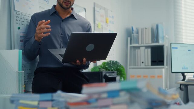 Thoughtful young Latin businessman stand with laptop analyzing data above paperwork stack. Hybrid work audit, digital transformation strategy, business corporate finance, executive decision concept.