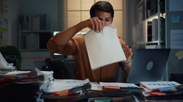 Overworked young Latin businessman working late on document in large stack of paperwork files at office night. Tax audit deadline, accounting stress, corporate burnout, urgent business concept.
