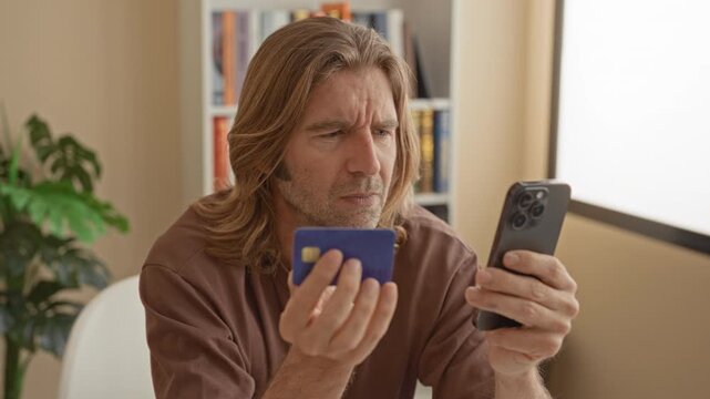 Man holding a creditcard and smartphone, frowning and squinting at the screen while seated in a studio with bookshelf and window light; frustration security concern.