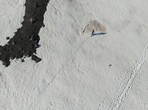 Aerial view of a lone hiker walking across a vast snowfield on the Versante sud dell' Etna next to dark volcanic rocks in Nicolosi, Sicilia, Italy.