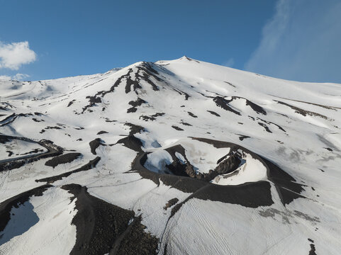 Aerial view of the snow-covered southern slope of Mount Etna featuring a volcanic crater and the majestic peak under a clear blue sky in Nicolosi, Sicilia, Italy.