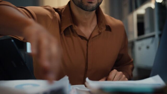 Close up overworked young Latin businessman working late on document in large stack of paper files at office night. Tax audit deadline, accounting stress, corporate burnout, urgent business concept.