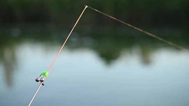 Fishing rod with alarm bell positioned above calm water, highlighting fishing line and soft blurred background. Perfect for representing outdoor leisure activity. evening fishing concept